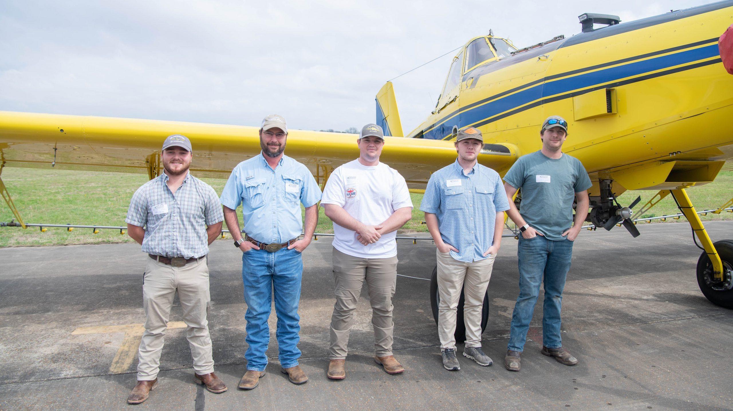 Five students standing in front of a crop duster plane on an airport runway.